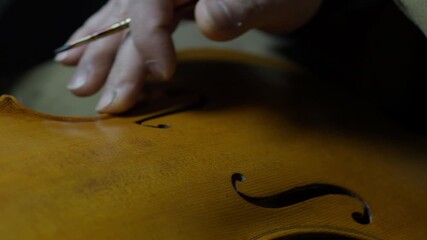 Hands of a luthier precisely darkening violin edges, simulating natural wear and aging on the handcrafted instrument on edges and purfling, with finger and paintbrush traditional tecqnique