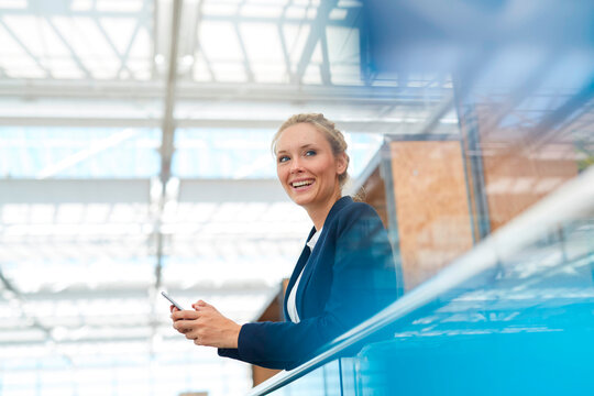Happy businesswoman leaning on railing with smart phone at airport