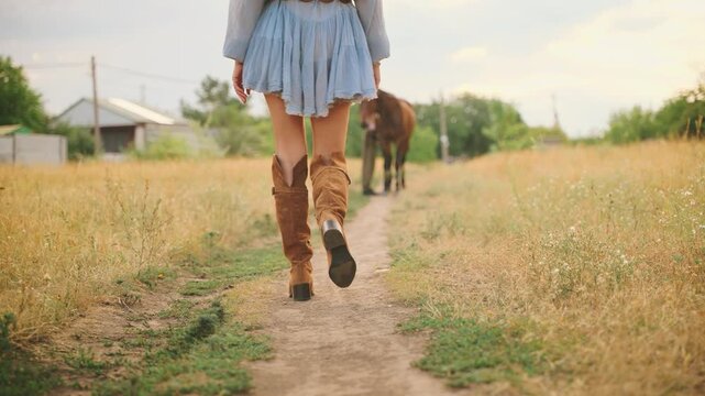 Woman with horse close-up body part legs in high leather brown boots walking along dirt country road green dry grass field summer nature. Sexy girl cowgirl shoes clothes wild west style back rear view