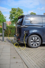 Electric minivan charges at a public EV station in an urban setting with a plugged yellow cable. Transition to eco-friendly mobility and the rise of green transportation in Europe’s cityscapes.