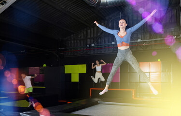 Positive middle-aged woman in sporty clothes jumping vertical high in modern trampoline hall