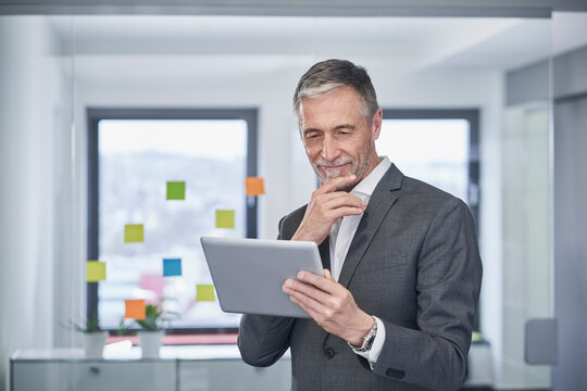 Smiling senior businessman with hand on chin using tablet PC in office