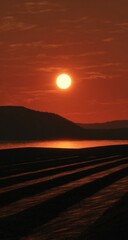 Red sunset over rows of ploughed fields, silhouetted against the glowing horizon. A hazy atmosphere obscures distant hills and casts a warm light on the water.