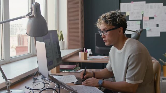 Portrait shot of young male UX designer drawing on graphics tablet then turning to camera with smile while working at desk with laptop and desktop computer at office, slow motion