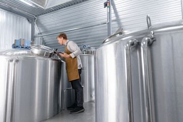 Worker caucasian male brewer inspecting stainless steel fermentation tanks in modern brewery with beer wort