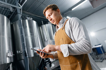 Caucasian male adult brewer using tablet in modern brewery with stainless steel tanks with craft beer