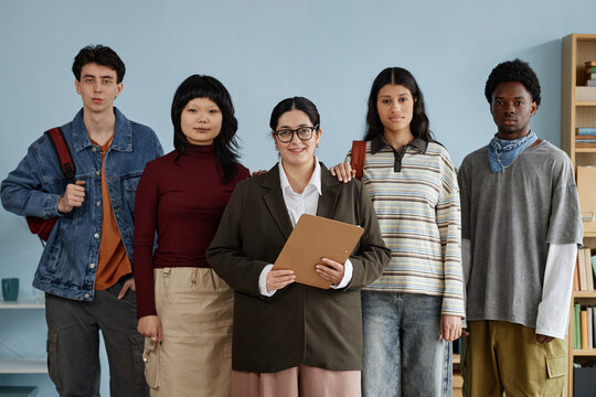 Group of multiethnic teenagers standing with female counselor holding clipboard during counseling session, teenagers posing together and looking at camera in educational setting