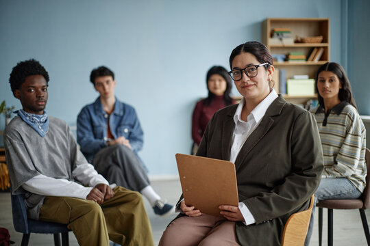 Multiethnic group of teenagers sitting in counseling session with woman counselor holding clipboard, teenagers listening attentively in therapy or support group setting