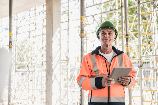 Thoughtful contractor holding tablet PC standing in front of scaffolding