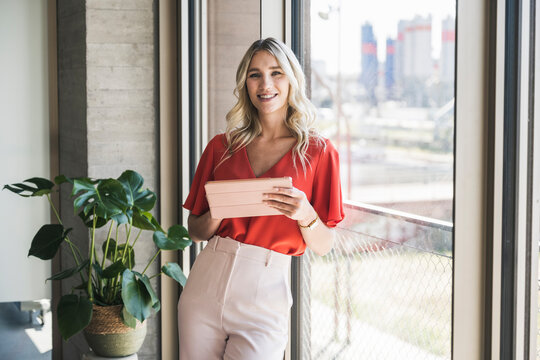 Smiling blond businesswoman with tablet PC leaning on window in office