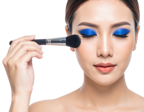 A woman with vibrant blue eyeshadow is applying makeup on her cheek using a large makeup brush with her eyes closed.