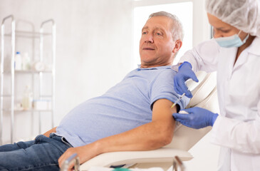 Obraz premium Old male patient lying on clinical chair having arm injection by middle-aged doctor