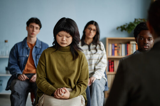 Group of multiethnic teenagers sitting in counseling session, Asian teenage girl with eyes closed and hands clasped in foreground, other teenagers listening in background