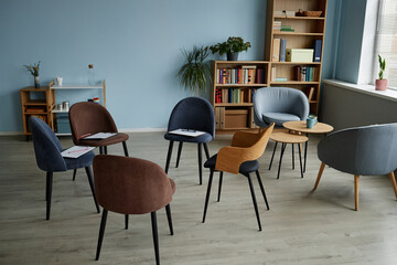 Empty counseling room with chairs arranged in circle and clipboards on seats prepared for group therapy session for teenagers, bookshelf and plants in background, sunlight coming through window