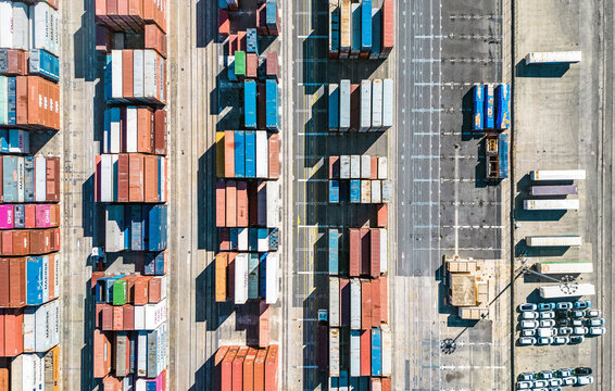 Aerial view of a tapestry of stacked shipping containers, arranged in neat rows, casting shadows on the concrete ground, creating a geometric pattern of industry, Valencia, Valencian Community, Spain.