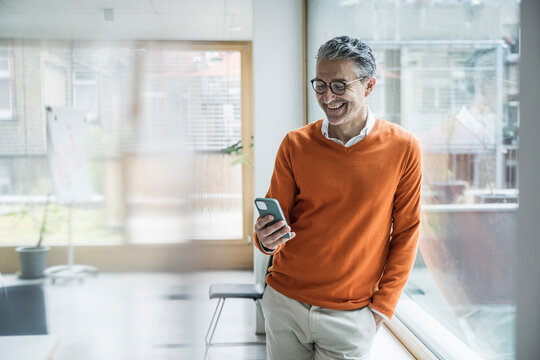 Smiling mature businessman using smart phone leaning at glass window in office