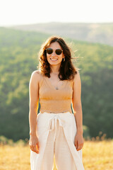 A joyful, happy woman wearing stylish sunglasses poses in a beautiful sunlit field, surrounded by lush green hills