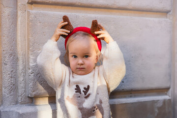 A young Caucasian girl with blonde hair wears a festive sweater and reindeer antlers. She poses against a textured wall, embodying the Christmas spirit.