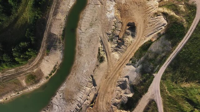 Aerial view of a sand pit where brown earth meets a dark green body of water, with a yellow excavator actively digging, Brussow, Brandenburg, Germany.