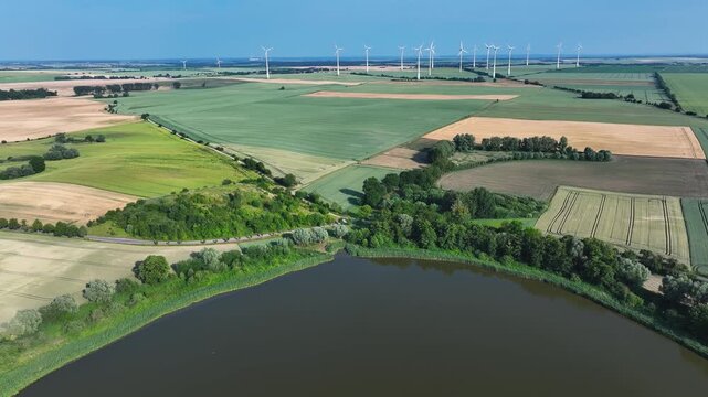 Aerial view of a serene lake bordered by lush trees and vibrant green fields, complemented by distant wind turbines under a clear sky, Brussow, Brandenburg, Germany.