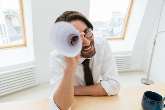 Smiling businessman looking through rolled up paper at office