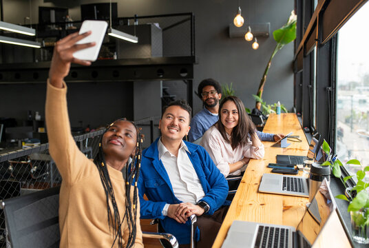Smiling businesswoman taking selfie through smart phone with colleagues in office