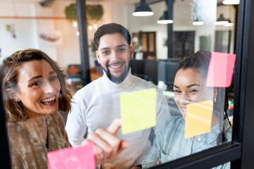 Smiling businesswoman with colleagues reading adhesive notes on glass in office