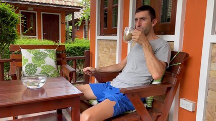 Man relaxing on bungalow porch, drinking coffee in the morning. Cozy tropical setting with wooden furniture, green plants, and peaceful holiday vibes on resort villa