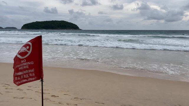 Red warning flag on tropical beach with &ldquo;No Swimming Here&rdquo; message in multiple languages, strong waves and cloudy sky in background, signaling dangerous sea conditions
