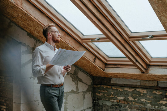 Thoughtful architect standing with blueprint looking through window at construction site