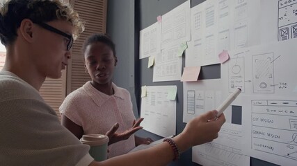 African American female UX designer developing user interface for digital product discussing mobile app wireframes with Asian male colleague pointing at paper plans hanging on wall in modern office - Powered by Adobe