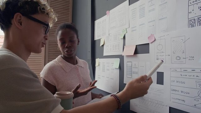 African American female UX designer developing user interface for digital product discussing mobile app wireframes with Asian male colleague pointing at paper plans hanging on wall in modern office