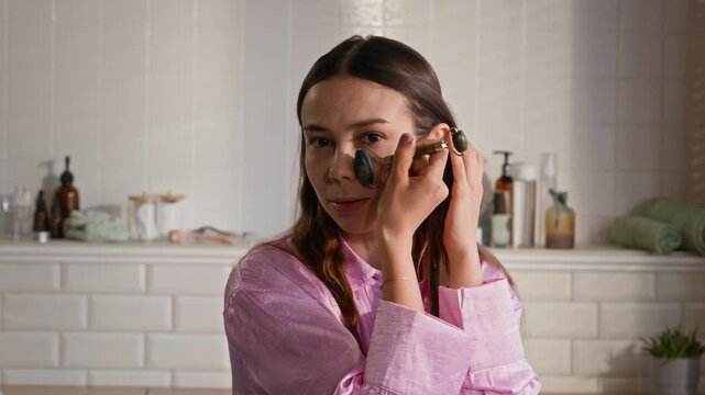 Young brunrtte woman using jade roller for massaging her face while standing in front of mirror in bathroom in the morning and having self care procedure
