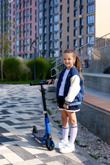 Little girl stands with scooter in front of multi-storey building background. School girl riding a scooter in city. Active leisure and outdoor sport for child. Eco-friendly urban transport for kid.