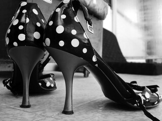 Black and white photo of vintage high heels with a polka dot pattern, viewed from a low angle on a tiled floor, creating a stylish and retro fashion aesthetic.
