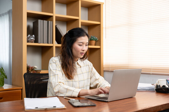 Asian woman working remotely at home, typing on laptop at desk with digital devices and paperwork, focused on task management and productivity in a cozy home office environment.