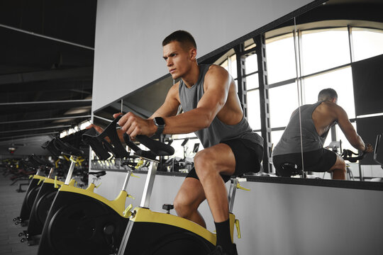 Handsome athlete young man in sportswear riding a stationary cycling bike in modern gym. Fitness and health care concept.