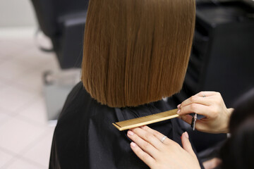 Naklejka premium Unrecognisable hairdresser cutting female client hair with professional barber scissors and comb and making stylish haircut short bob hairstyle in beauty salon. Hair care, hair salon and hairstyling