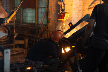 Workers at metallurgical plant pour molten metal from furnace into foundry ladle