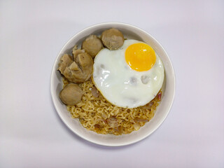 A white bowl filled with instant noodles, garnished with a fried egg, and various meatballs, all set against a plain white background. Represent food, Asian cuisine, comfort food, quick meal