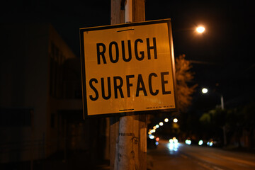 Obraz premium Yellow and black rough surface traffic sign, attached to a power pole, beside a road at night