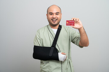 An Asian man in a hospital gown smiles gently at the camera with his arm bandaged and bandaged, posing holding a health insurance card to demonstrate resilience and positive attitude during recovery.