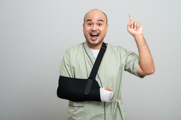 Asian man in patient attire smiles gently at the camera, wearing an arm sling and bandage, striking...