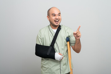 Asian man in hospital gown smiles gently at camera with arm bandages and bandages and crutches, posing to show resilience and positive attitude during recovery.