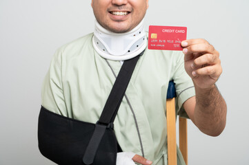 Asian man in hospital gown smiles gently at camera with arm bandages and bandages and crutches, posing holding health insurance card to demonstrate resilience and positive attitude during recovery.