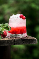 Chilled raspberry cocktail with ice cubes placed outdoors against natural background of lush raspberry bushes. Summer drink concept