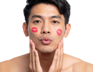 A young man with lipstick kiss marks on his cheeks poses with puckered lips and hands gently touching his face against a black background.