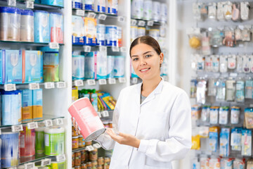 At pharmacy, friendly female apothecary holds jar of baby formula in hands and talks about properties of product. Visitor shopping in background