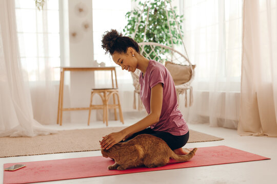 Young woman in sportswear enjoying cozy moment at home with ginger cat while sitting on a yoga mat