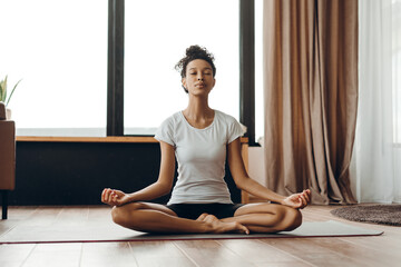 Young African American woman meditating on yoga mat in lotus pose at home